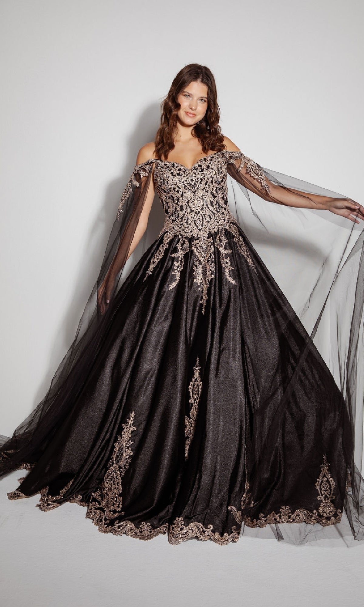 A woman wears the Quinceanera Dress EK1002 by Eureka—a black, off-the-shoulder gown with gold embroidery, sheer tulle sleeves, and a lace-up back—standing against a plain background.